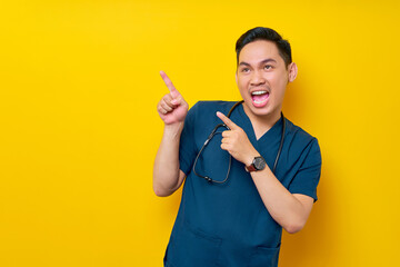 Smiling professional young Asian male doctor or nurse wearing a blue uniform standing confident while pointing a finger at copy space isolated on yellow background. Healthcare medicine concept