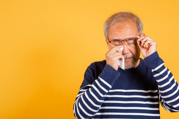 Asian elder man crying raise glasses with tissue wipe red eyes studio shot isolated on yellow background, Portrait senior old man sad wiping away his tears, Upset depressed lonely