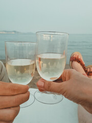 female and male hands hold glasses of white wine on a sandy beach against the backdrop of the sea. summer vacation and beach holiday concept. soft focus. vertical photo