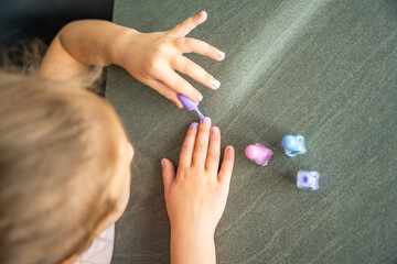 Top view of little girl doing manicure and painting nails with colorful pink, blue and purple nail polish at home.