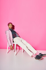 young handsome man in pink blazer with white pants sitting on chair on pink backdrop, doll like