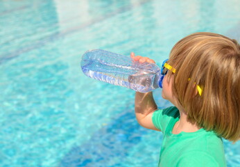 Cute little boy drinks water from a plastic bottle against the background of a swimming pool....