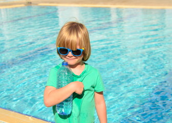 Cute little boy drinks water from a plastic bottle against the background of a swimming pool....
