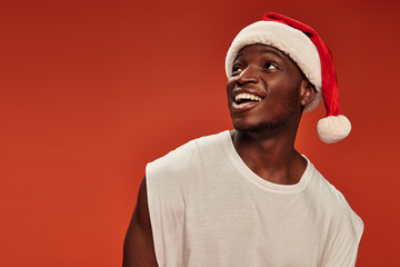 amazed and overjoyed african american man in santa cap smiling and looking away on red backdrop - obrazy, fototapety, plakaty