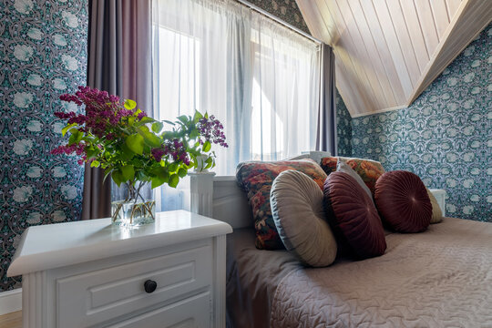 White bed in bedroom, bedside table with standing bouquet of lilacs