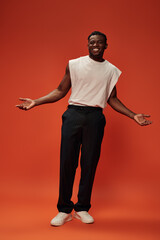 Fototapeta na wymiar happy african american man in trendy casual clothes showing welcoming gesture with open arms on red