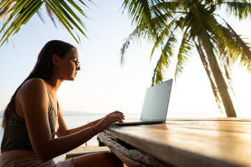 Freelancer girl with a computer among tropical palm trees work on the island in sunset