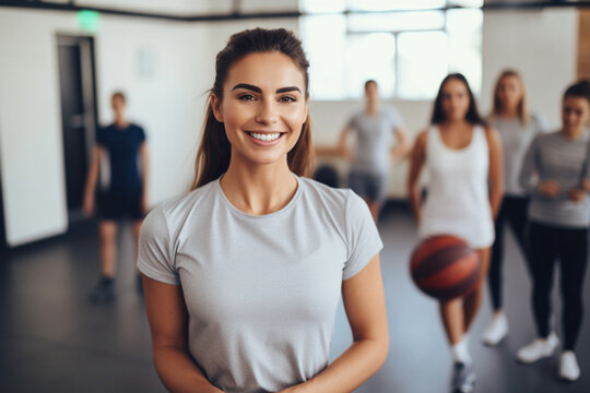 Portrait of Young beautiful female basketball coach standing in school gym , holding basketball ball , looking at camera and smiling
