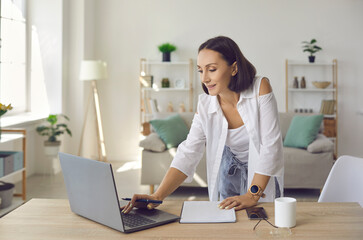 Woman working on computer in home office. European businesswoman in 30s 40s standing by desk with laptop, phone and coffee cup, replying to business emails, visiting website, taking notes in organizer