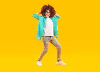 Fototapeta na wymiar Happy funny preteen boy in curly wig dancing and having fun. Full size photo of joyful boy wearing glasses, turquoise shirt, jeans and sneakers standing against yellow studio background