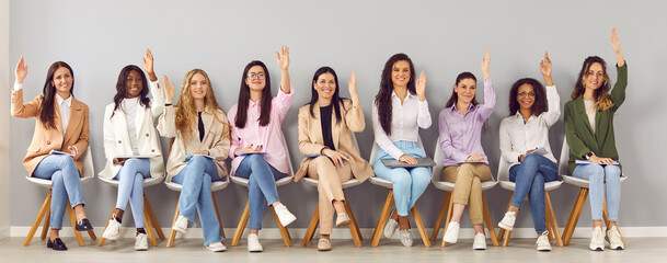 Portrait of group of happy diverse business women company employees looking at camera and raising hands to ask or answer a question sitting in a row on meeting on a gray wall background. Banner. - obrazy, fototapety, plakaty