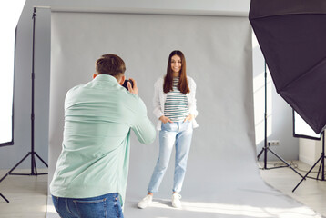 Back view portrait of a young man photographer shooting girl by digital camera in his hands. Beautiful young female model posing in modern professional photo studio with light equipment.