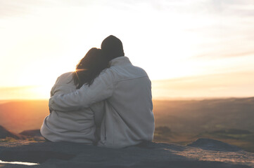 Back view of the happy couple in love sitting on top of a mountain enjoying a sunset landscape view toned image