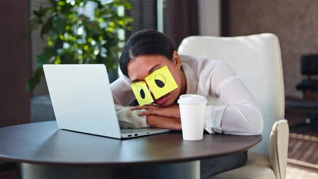 Exhausted african american woman in formal clothes sleeping on office desk with yellow stickers on eyes. Lazy unproductive female office worker leaning head on hands near opened wireless laptop.