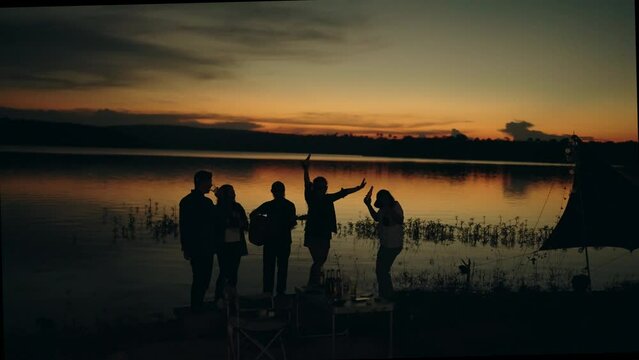 Group of friends playing guitar and drinking beer on silhouette sunset background beautiful nature, mountains and lake,Friendship and celebration concept.