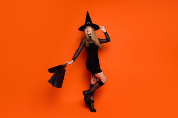 Full length photo of dreamy pretty girl dressed dark witch dress headwear holding bargains empty space isolated orange color background