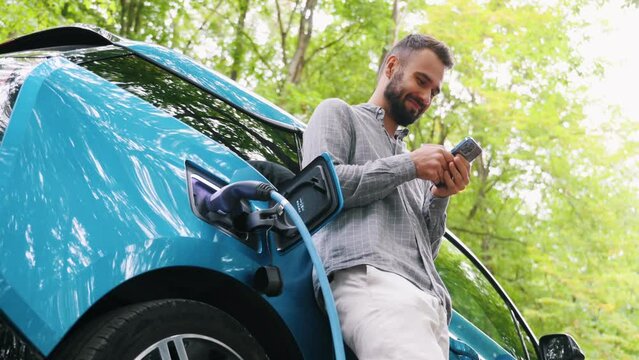 Man stands with phone near her electric car and waits when vehicle will charged at green forest. Save ecology alternative energy sustainable of future.