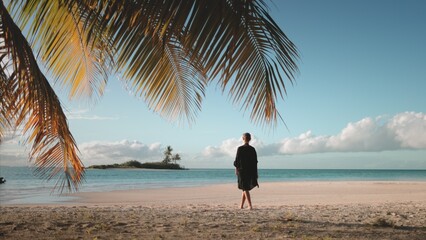 Woman walking barefoot white sand beach at sunset. Tourist girl in black dress enjoy tropical...