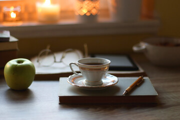 Vintage books, e-reader, pen, reading glasses, phone, cup of tea or coffee, bowl of cookies, green apple and lit candles on the table. Hygge at home, studying atmosphere. Selective focus.