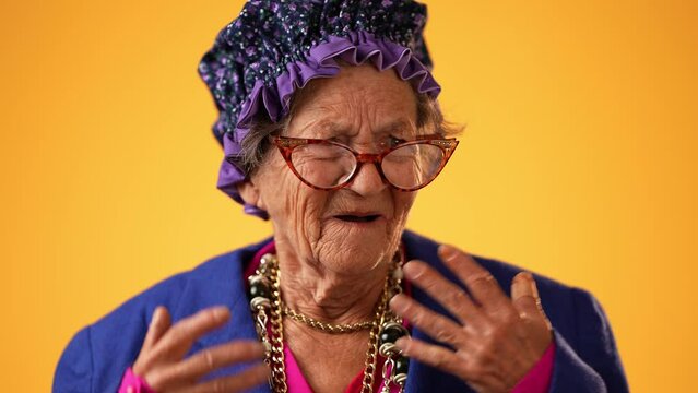 Closeup of angry, scared elderly senior old funny crazy woman with wrinkled skin and grey hair put hands out ask WHY ME, isolated on yellow background studio. People emotions