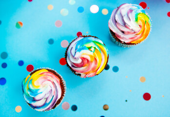 Diagonal line of three rainbow colors cupcake on blue background with glitter confetti from top view