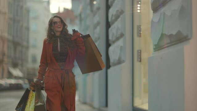 Hello november. happy trendy 40 years old woman in brown trench coat with shopping bags and autumn yellow leaves in the city.
