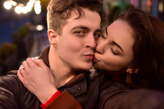A young couple in love walks down a night street and takes selfies. The girl is kissing her boyfriend.