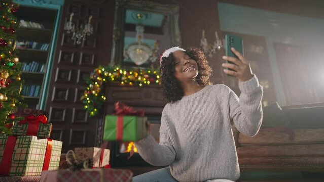African American woman laughing posing on Christmas tree background at home blogger shooting vlog. Call friends and family for christmas wishes. Girl sit near christmas tree make a video call online