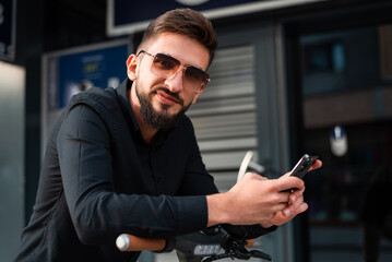 A young urban stylish man with electric scooter standing in front of modern business building on city street and using a phone.