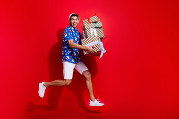 Fototapeta na wymiar Full body photo of handsome young guy running jump gifts pile wear santa claus print x-mas outfit isolated on red color background