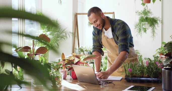 African American man flower shop manager standing in apron in small floral shop. Businessman working in flower shop writing concentrated on business planning. Floristry business and retail concept.