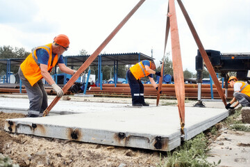 Builders in hard hats work on construction site. Team of workers lays concrete slab on ground on...