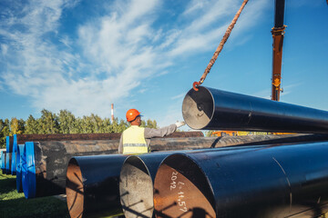 Slinger unloads large gasification pipes on summer day. Pipes for transporting natural gas. Construction of gas pipeline. Storage of large diameter metal pipes. - obrazy, fototapety, plakaty
