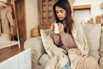 Woman freezes in winter time. Young woman with plaid near electric heater, holding a cup of hot...