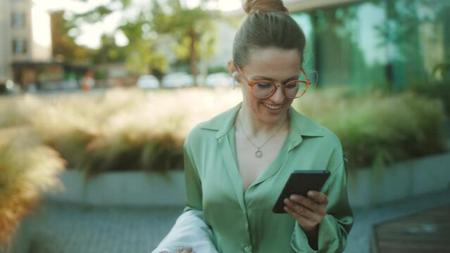 smiling modern woman worker near business center in green blouse and eyeglasses with briefcase and wireless headphones using smartphone.