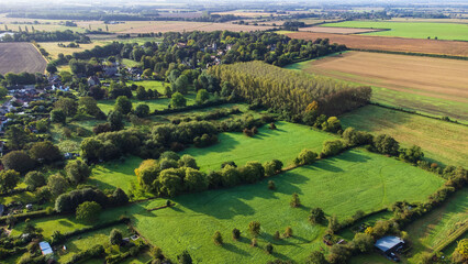View of fields over a village in Oxfordshire. Trees surround each of the grassy fields. In the background plowed fields.