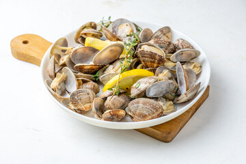 White plate with cooked vongole clams with lemon and herbs, on a white table on a board close-up.