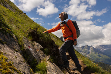 Sporty man with backpack standing on steep trail against bright cloudy sky and looking up, Austria - obrazy, fototapety, plakaty