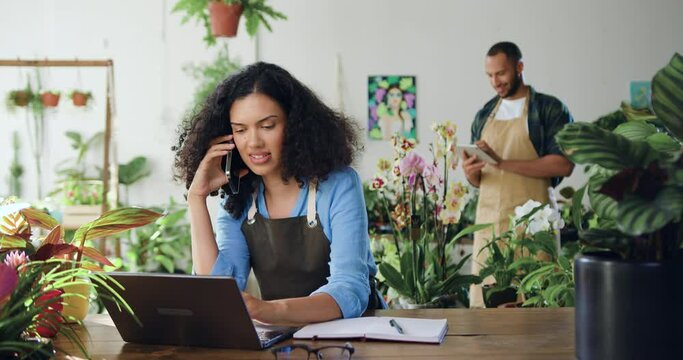 African american woman in apron sitting on desk speaking to client on mobile phone and using laptop man working which digital tablet in background. Family florists using laptop, typing and talking