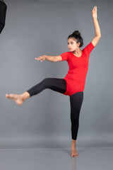 Healthy young woman in red t-shirt and black leggings doing yoga and stretching exercises isolated on grey background