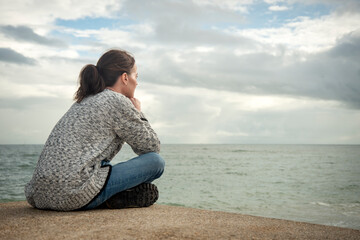 Woman sitting alone looking out to sea deep in thought, rear view, Autumn.