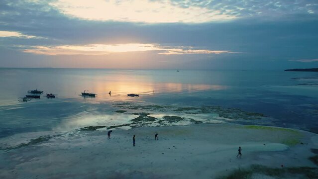 Filipino teenagers playing football on Tubod beach, Siquijor island, Philippines. Fisherman in the sea. Cinematic aerial drone view at sunset. Vibrant cloudy sky colors. Famous travel destination.