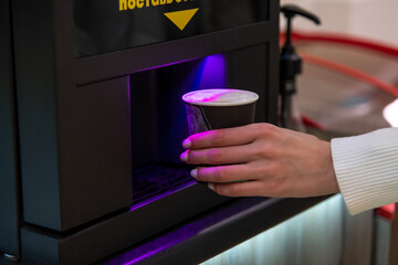 Close-up view of female hand taking out black disposable paper coffee cup with latte coffee drink from table top vending coffee machine in micro market. Soft focus. Self service theme.