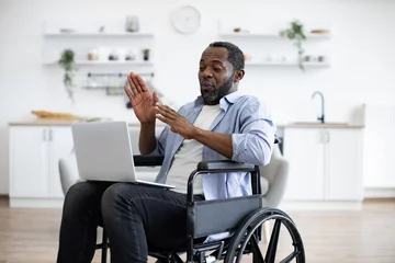 Fotobehang Young african american person in casual wear with portable computer while sitting in wheelchair in studio apartment. Positive adult man having video call, while sitting at kitchen. © sofiko14