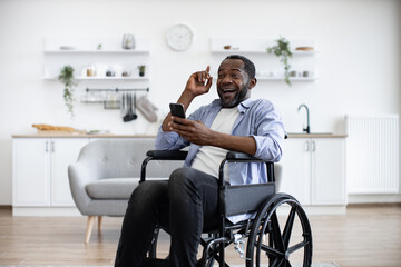 Young african american person in casual wear with smartphone sitting in wheelchair in studio apartment. Positive adult man pointing with finger up while inspired by great idea.
