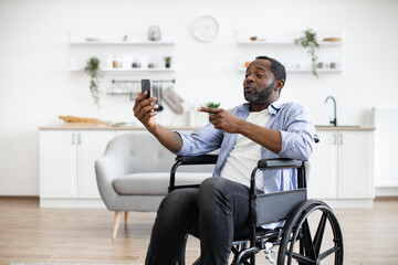 Cheerful wheelchair user in casual outfit utilizing phone webcam while enjoying free time indoors....