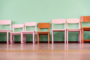Several wooden children chairs of different sizes stand in row along empty wall on floor of classroom - obrazy, fototapety, plakaty