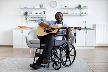 Talented african musician sitting in wheelchair holding acoustic guitar in bright dining room....