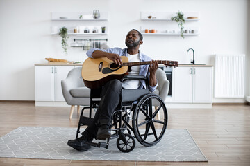 Talented african musician sitting in wheelchair holding acoustic guitar in bright dining room....