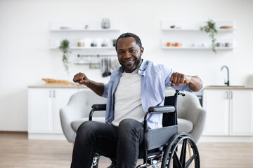 Close up of joyous bearded man in wheelchair exercising stretching arms while following workout program at home. Positive african adult in cozy outfit participating in physical activity in apartment.